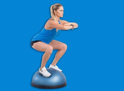 Side view portrait of sporty attractive woman wearing black shorts and blue top working in gym doing exercise in bosu balance trainer, squats on fitness ball, holding balance. Indoor studio shot.
