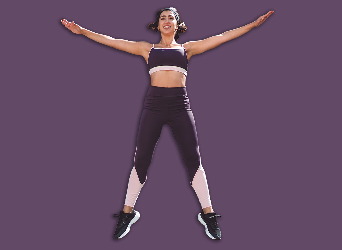 Fit woman in perfect jumping jack pose, mid-air with arms and legs extended, capturing alignment, control, and form in a synchronized cardio movement at peak extension.