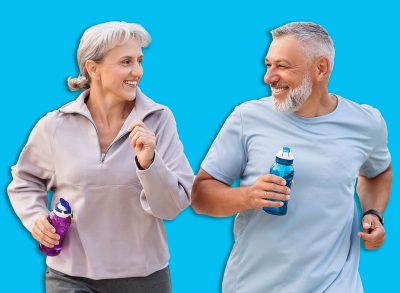 Lovely joyful retirees couple jogging outside in city park along alley with green trees, happy husband and wife looking at each other with smile holding water bottles in hands