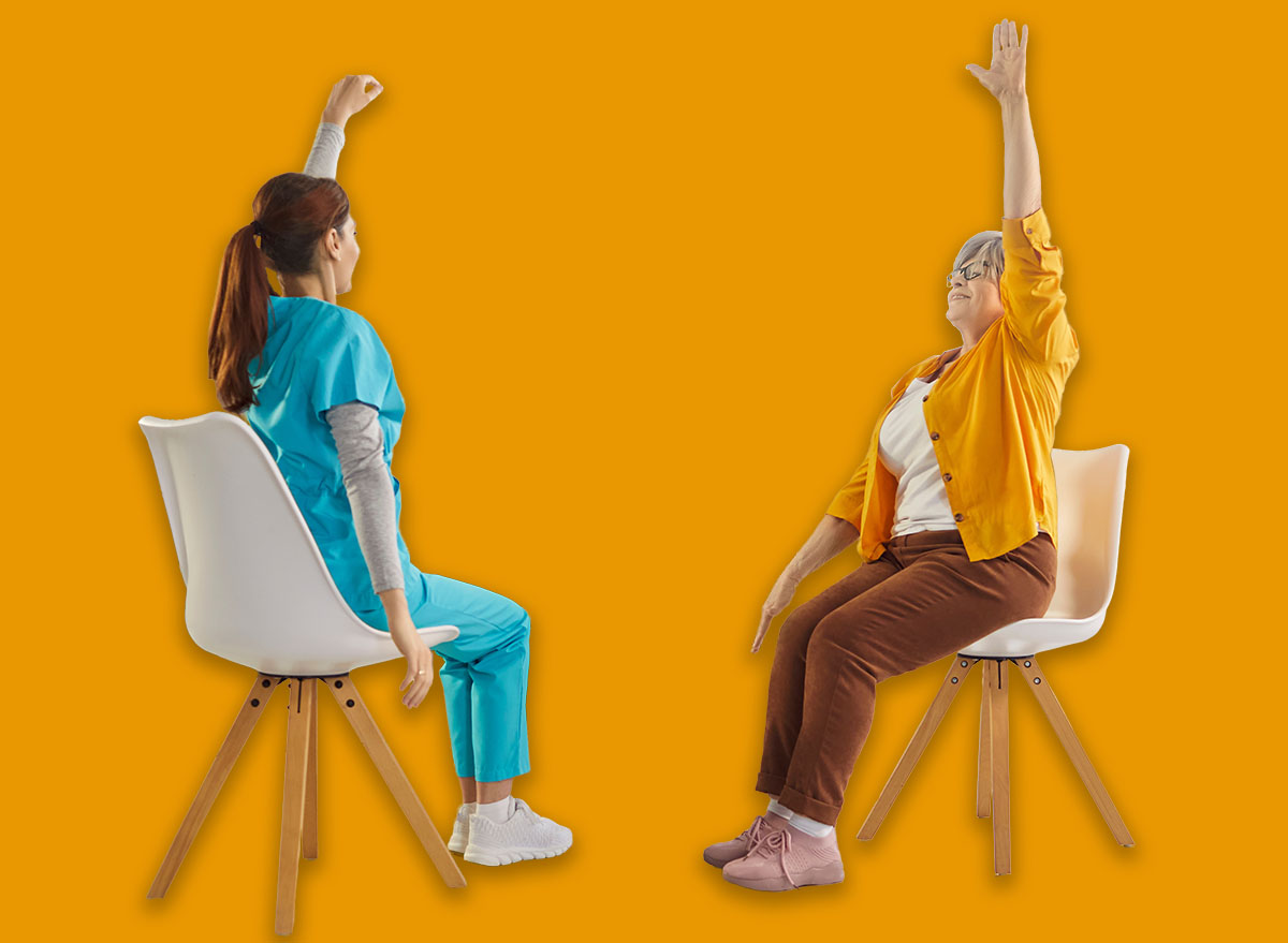 Young nurse leading seated exercise session with elderly patients in nursing home. Group of happy senior people sitting on chairs, raising their arms during physical therapy for mobility and wellness.
