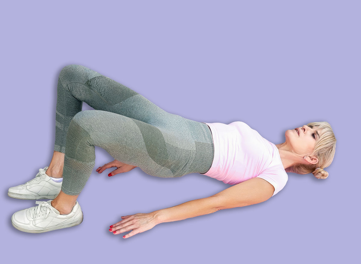 Young woman lying on back on pink exercise mat outdoors performing glute bridge with resistance band around thighs, hips raised to engage glute and hamstring muscles, wearing athletic leggings and
