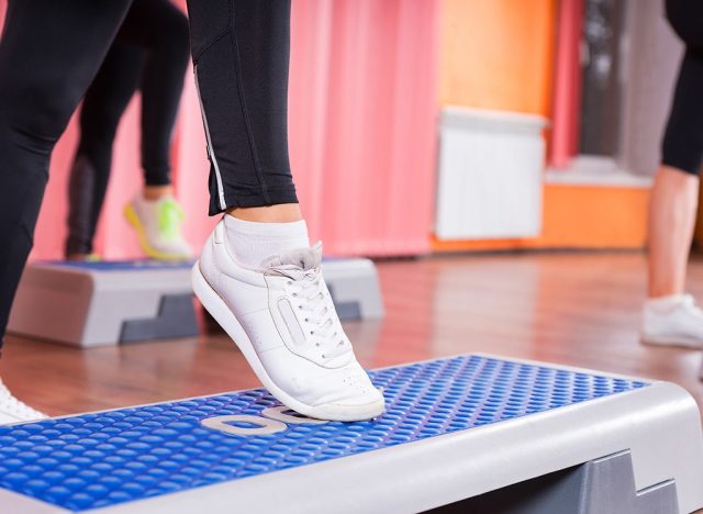 Close Up of Woman Wearing White Sneakers Doing Toe Tap on Step Platform in Aerobic Exercise Class with Group of Women in Background in Dance Studio