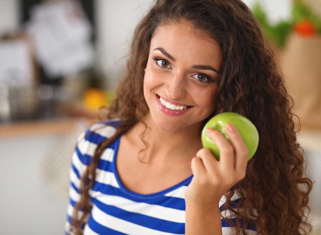 menina com cabelos longos sorrindo, segurando uma maçã verde